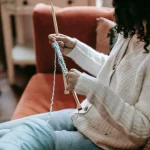 Side view of crop anonymous female artisan with yarn and needles knitting on couch in living room